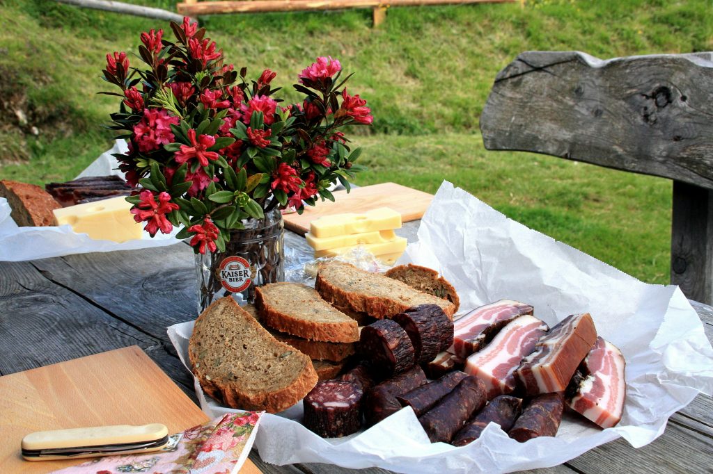 Planche de charcuterie, pain et fromage disposés sur une table en bois à l’extérieur, avec un bouquet de fleurs dans une chope de bière Kaiser.